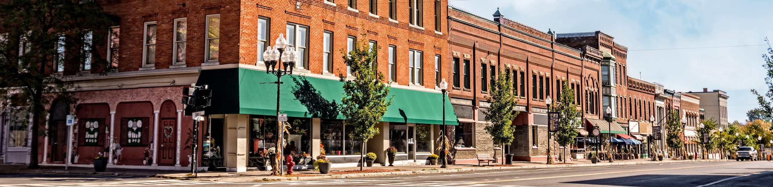 Modern bank building exterior at dusk with warm interior lights visible through large windows. Cars parked in lot, tree nearby
